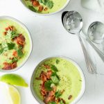 Overhead shot of Bacon Avocado Soup in three white bowls with three silver spoons and two quarter sliced lemons laying beside on a white background.