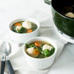 Image of Keto Low Carb Italian Wedding Soup in two white bowls with black handle spoons and a balck pot on either side laying on a white tea towel.