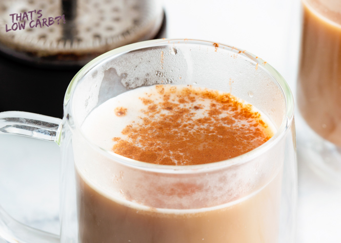 Image of Low Carb Keto Pumpkin Spice Latte in a clear glass mug with pumpkin spice sprinkled on top, with blurred second mug and french press in the background.