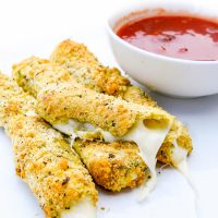 Image of Low Carb, Keto Mozzarella Sticks with one in half and cheese oozing out and red dipping sauce in a small white bowl beside.