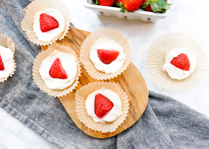 Image of keto cheesecake fat bombs with strawberries on top on a wood cutting board and a dark grey tea towel.