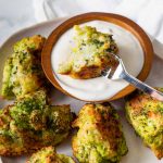 Image of Keto Broccoli Tots on a white plate with one bitten in half being dipped in white sauce in a brown bowl .