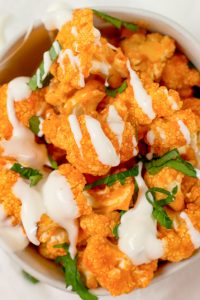Overhead shot of Keto Buffalo Cauliflower Bites in a white bowl with white sauce drizzled over top and green garnish on a white background.
