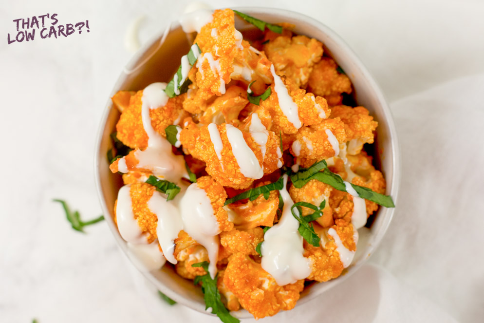Overhead shot of Keto Buffalo Cauliflower Bites in a white bowl with white sauce drizzled over top and green garnish on a white background.