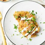 Overhead shot of keto Parmesan chicken with white sauce drizzled on top and green garnish sprinkled over on a white plate. Bronze fork next to plate and a bowl of keto low carb breadcrumbs in a glass bowl on w white background.
