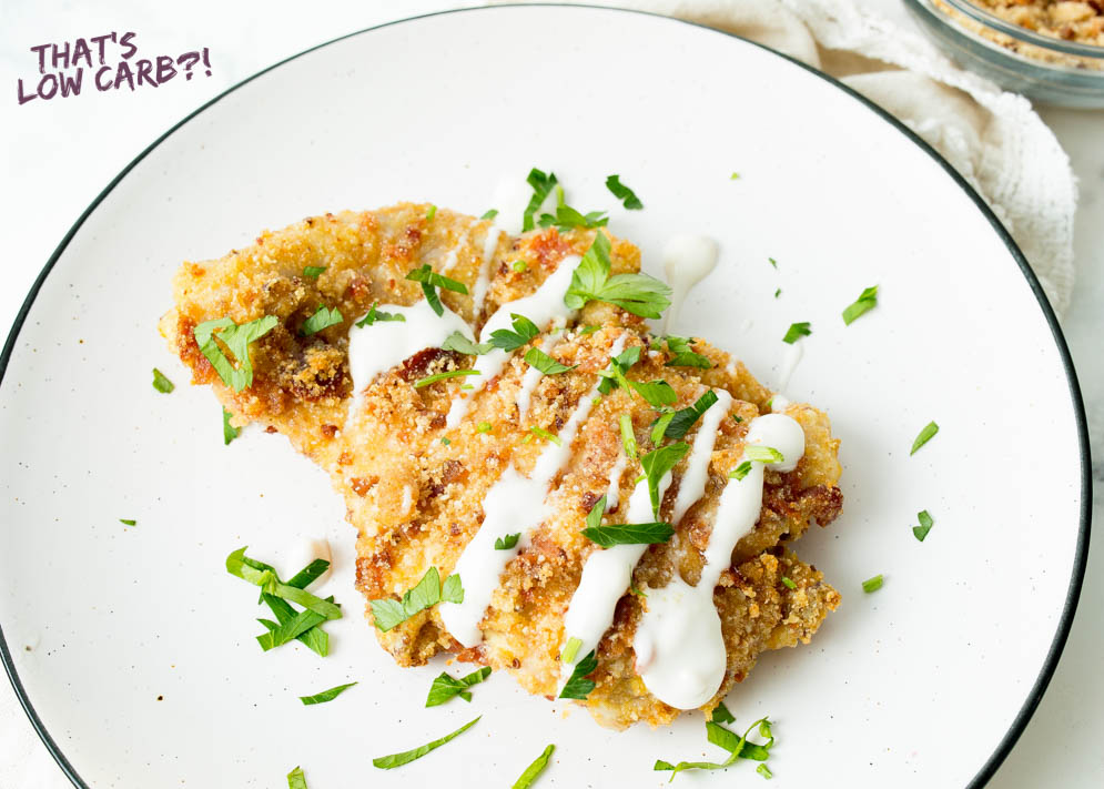 Overhead shot of keto parmesan chicken with white sauce drizzeled over top and green garnish sprinkled around white plate.