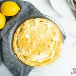 Overhead shot of baked sugar-free lemon meringue pie with dark blue cloth and lemons to the left.