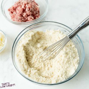 Biscuits batter mixed with a whisk and a glass bowl of uncooked sausage beside